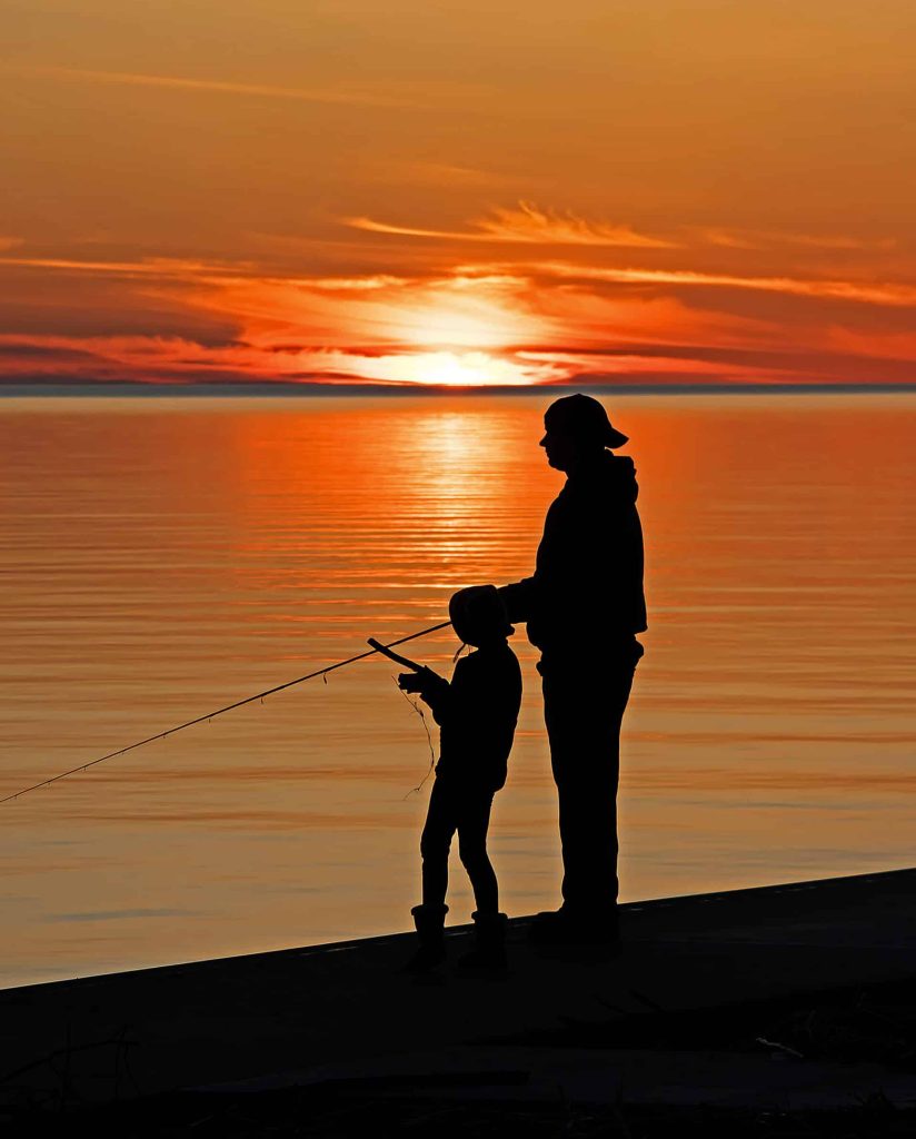 Silhouettes of an adult and child fishing on a Lake Michigan beach near Glen Arbor at sunset, with the orange and red sky reflected on the water.