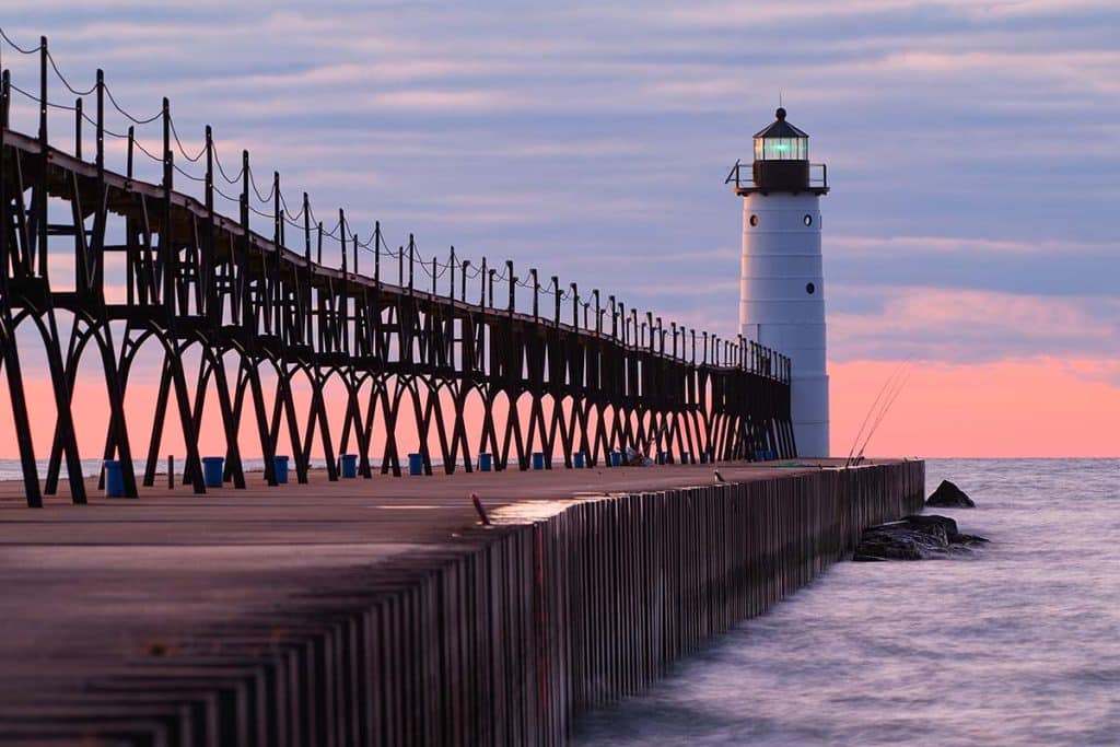 The Manistee North Pierhead Lighthouse at the end of its catwalk pier, photographed at sunset with a pink and blue sky.