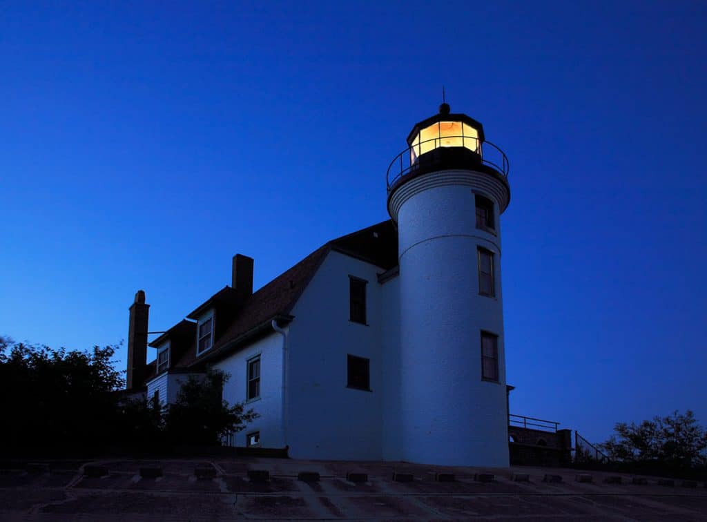 Point Betsie Lighthouse lit at dusk, with the lantern room glowing against the deep blue evening sky.