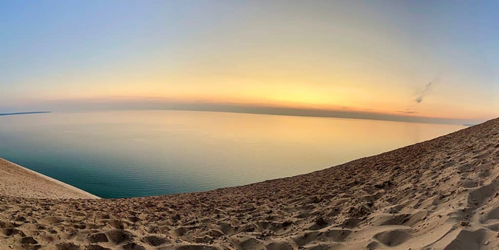 Panoramic view of Lake Michigan from the top of a sand dune at Sleeping Bear Dunes National Lakeshore, with an orange sunset sky.